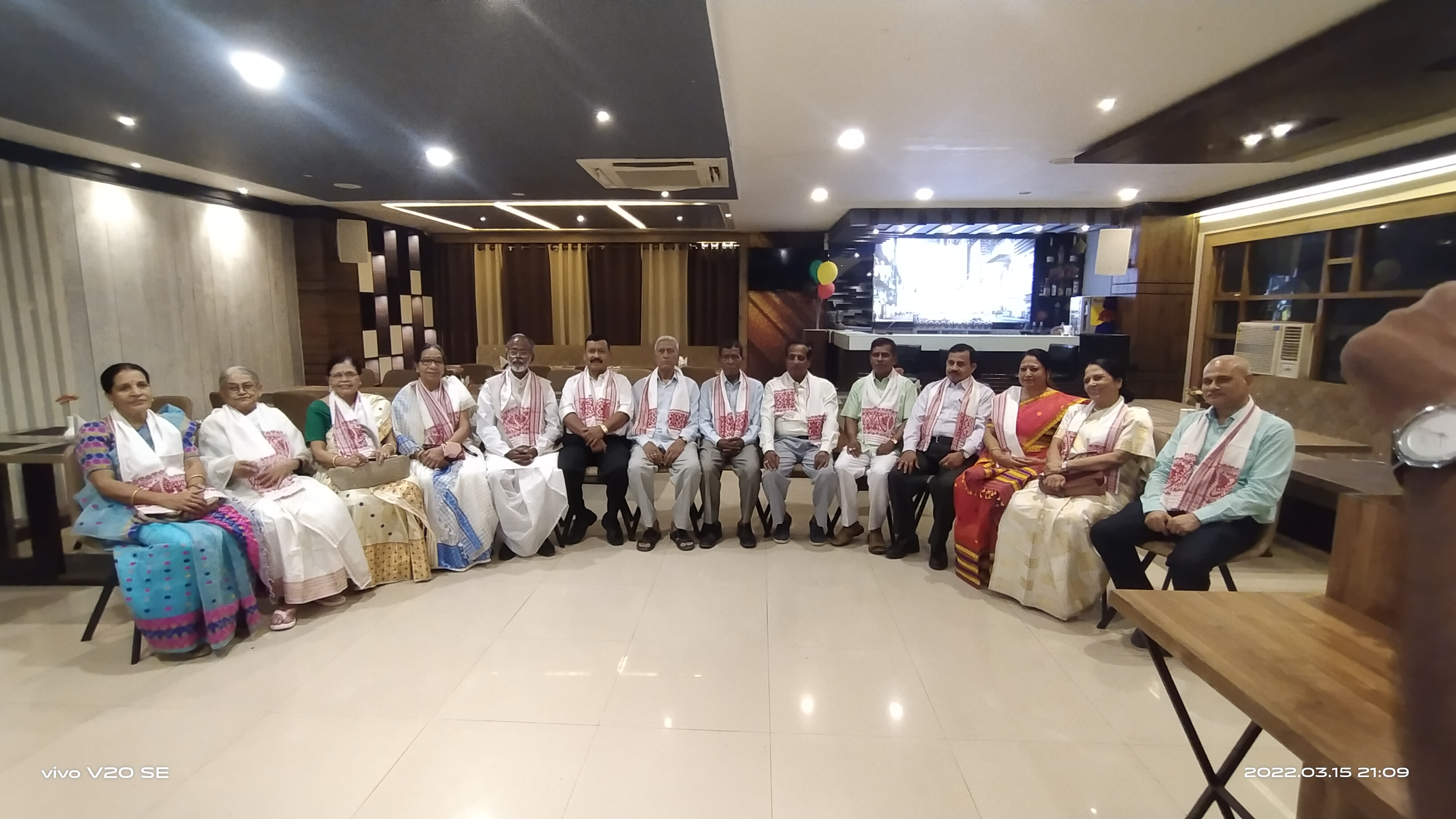 Alumni members seated together indoors during an association gathering, many wearing traditional Assamese gamusa scarves.