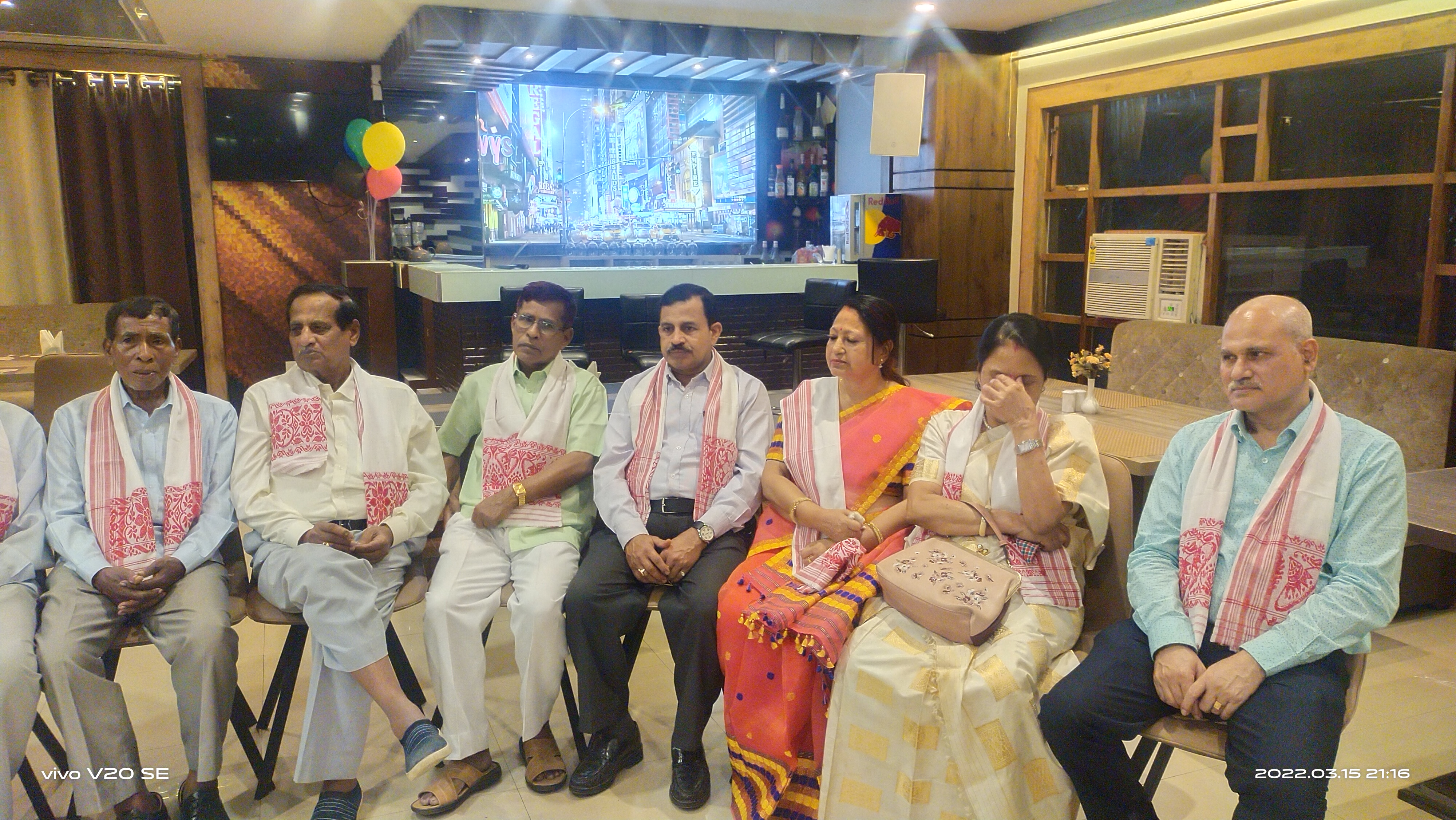 Alumni attendees seated indoors during an association event, with traditional Assamese scarves and formal attire.