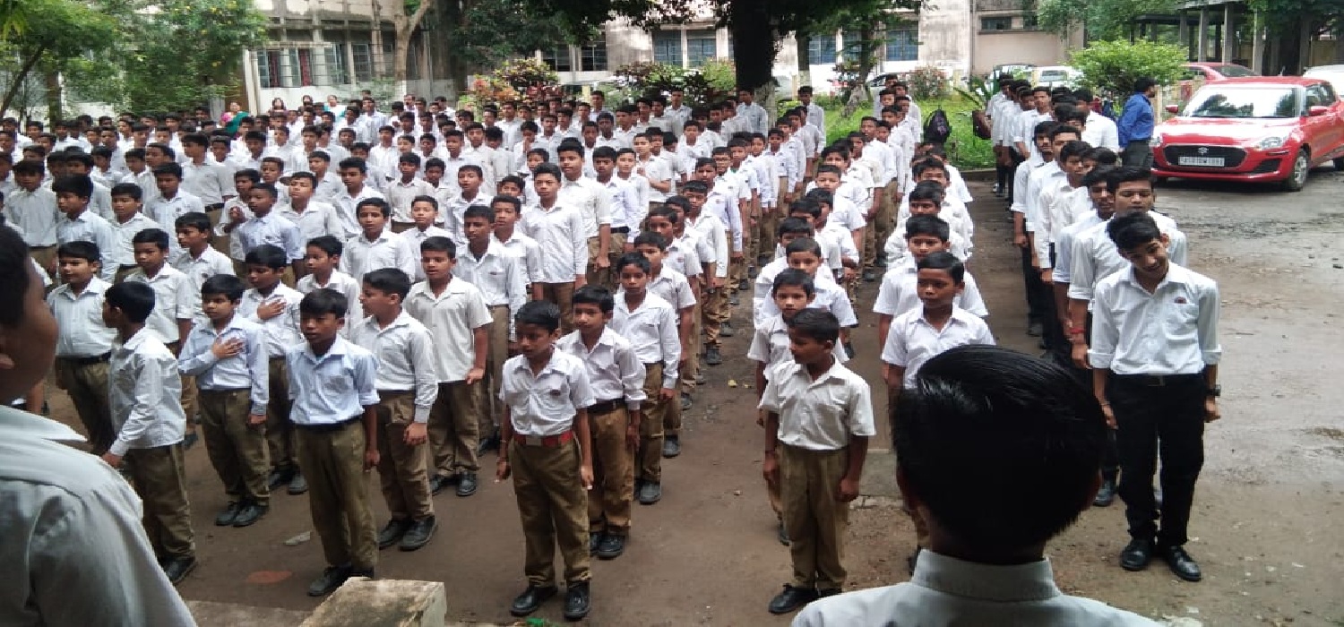 Students in uniform standing in rows during a school assembly in a courtyard, with a building and trees in the background and a person addressing them from the front.