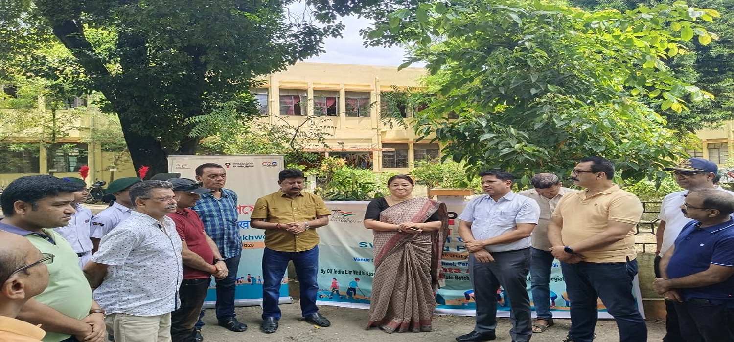 Group of adults standing in a school courtyard near a banner related to a cleanliness or awareness initiative, with trees and a school building in the background.