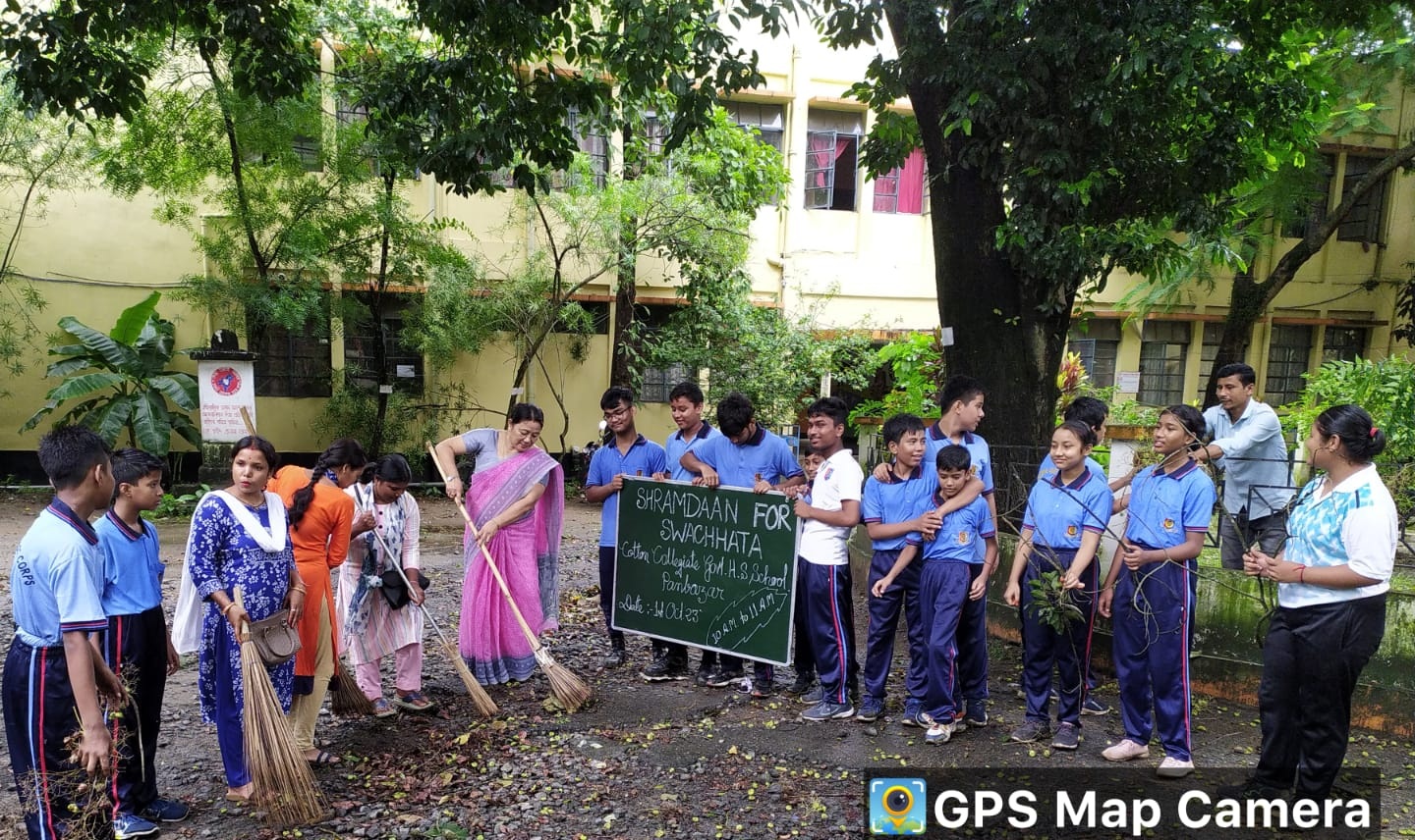 Students and staff participating in a cleanliness drive at a school courtyard, holding tools and a signboard promoting “Shramdaan for Swachhata,” with trees and a school building in the background.