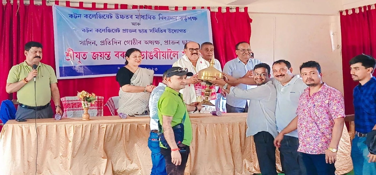 Group of adults on a stage holding a ceremonial lamp during an indoor school event, with a banner and red curtains in the background and a speaker addressing the audience.